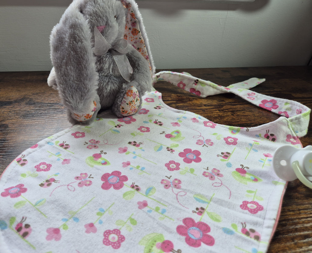 Nom Nom ABDL bib in white floral and ladybird print laid flat on wooden surface showing full bib shape with contrast hem