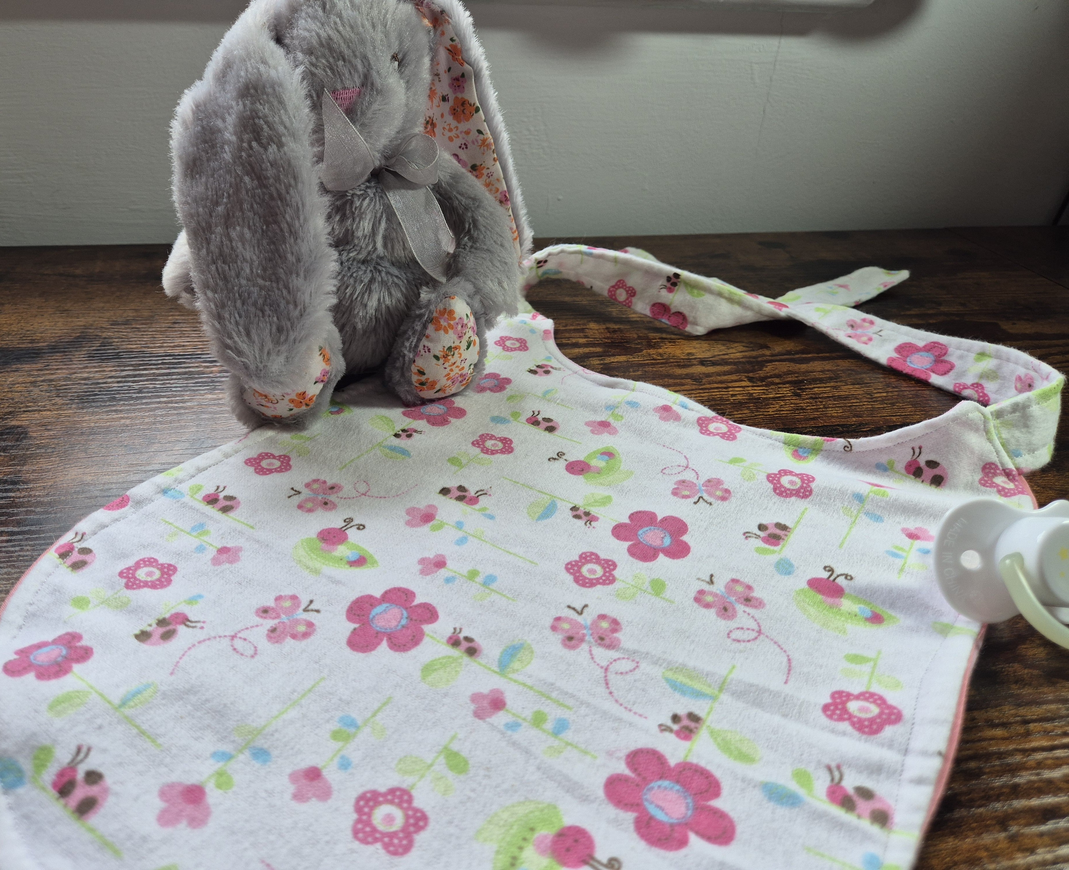 Nom Nom ABDL bib in white floral and ladybird print laid flat on wooden surface showing full bib shape with contrast hem