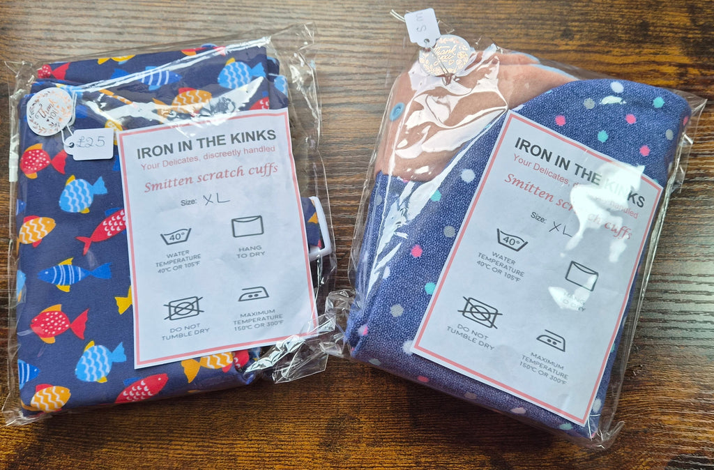 Two packaged cotton smitten scratch cuffs on a wooden table. The left cuff is navy blue with colorful fish patterns, and the right cuff is dark blue with white polka dots.