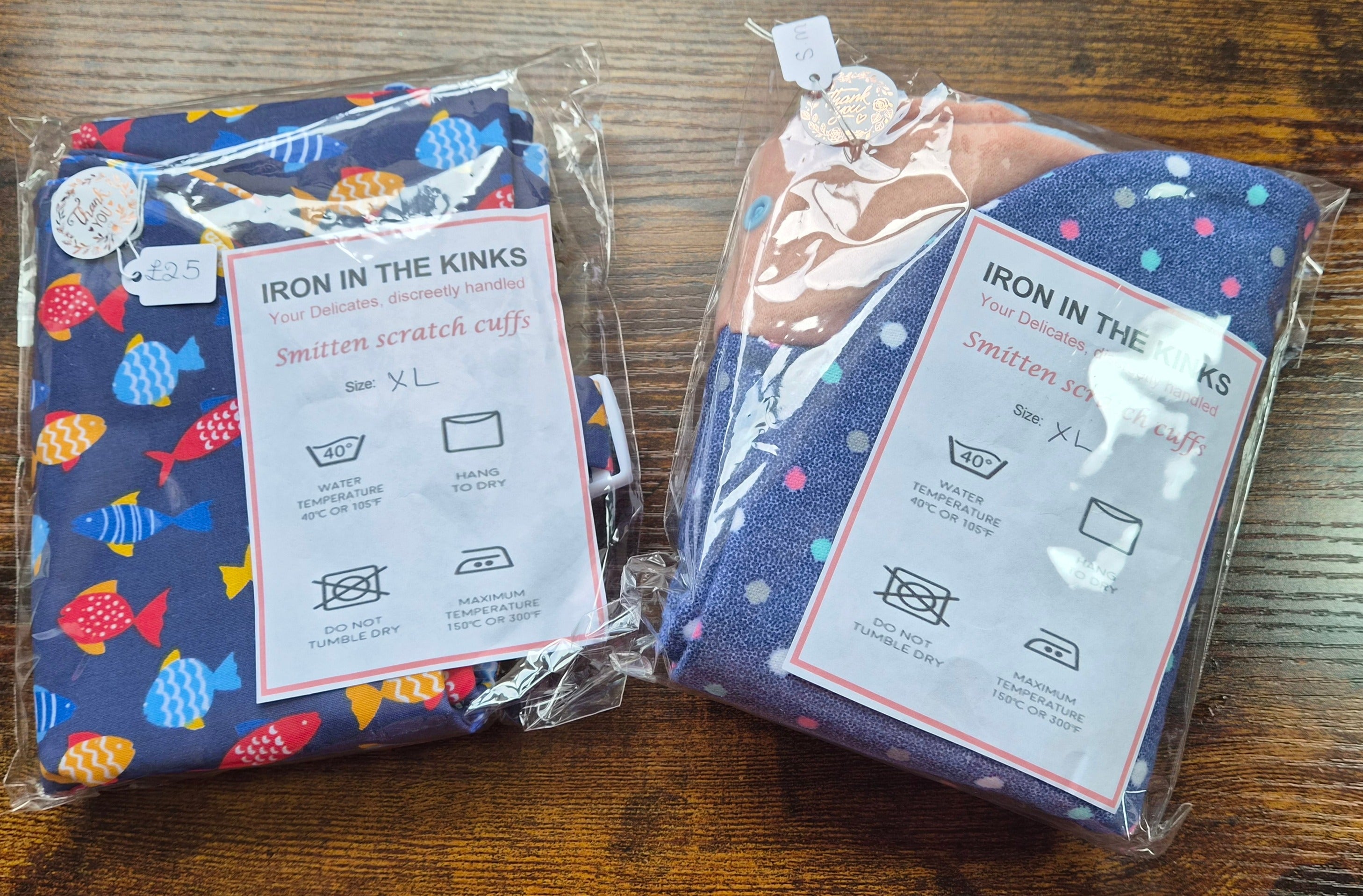 Two packaged cotton smitten scratch cuffs on a wooden table. The left cuff is navy blue with colorful fish patterns, and the right cuff is dark blue with white polka dots.