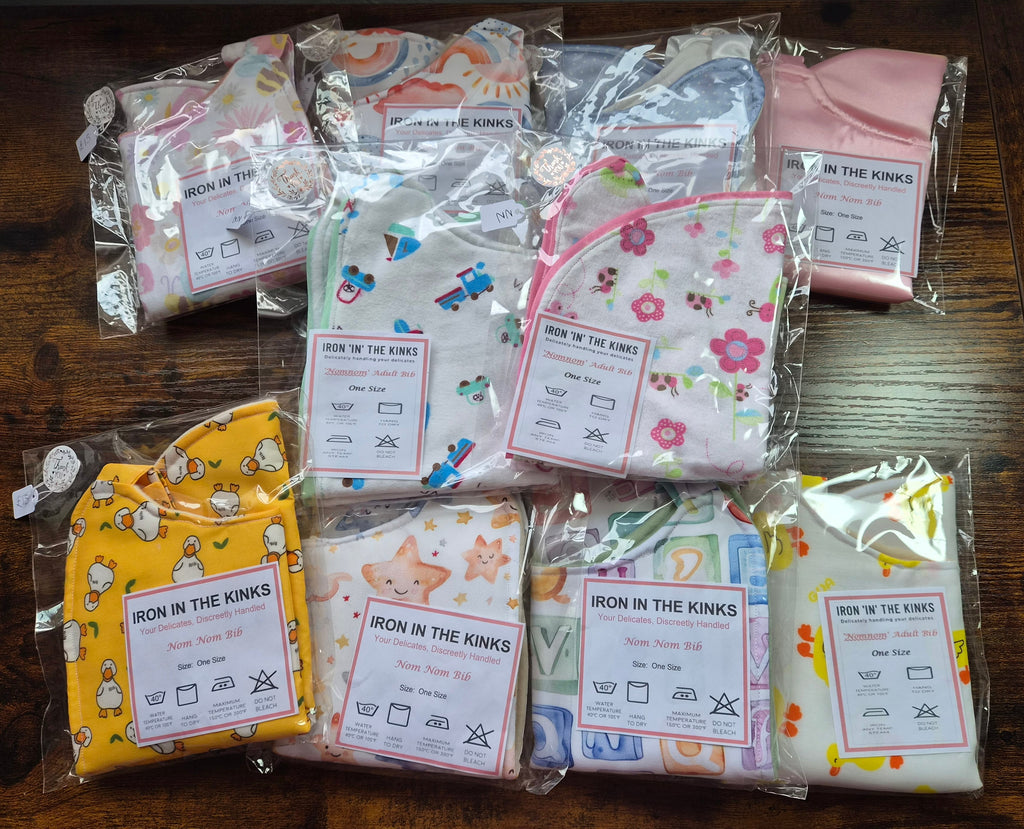 Seven Iron in the Kinks Nom Nom Bibs in assorted prints — rainbow, trains, floral, ducks, stars, alphabet blocks and fruit — in clear packaging with care labels, one size