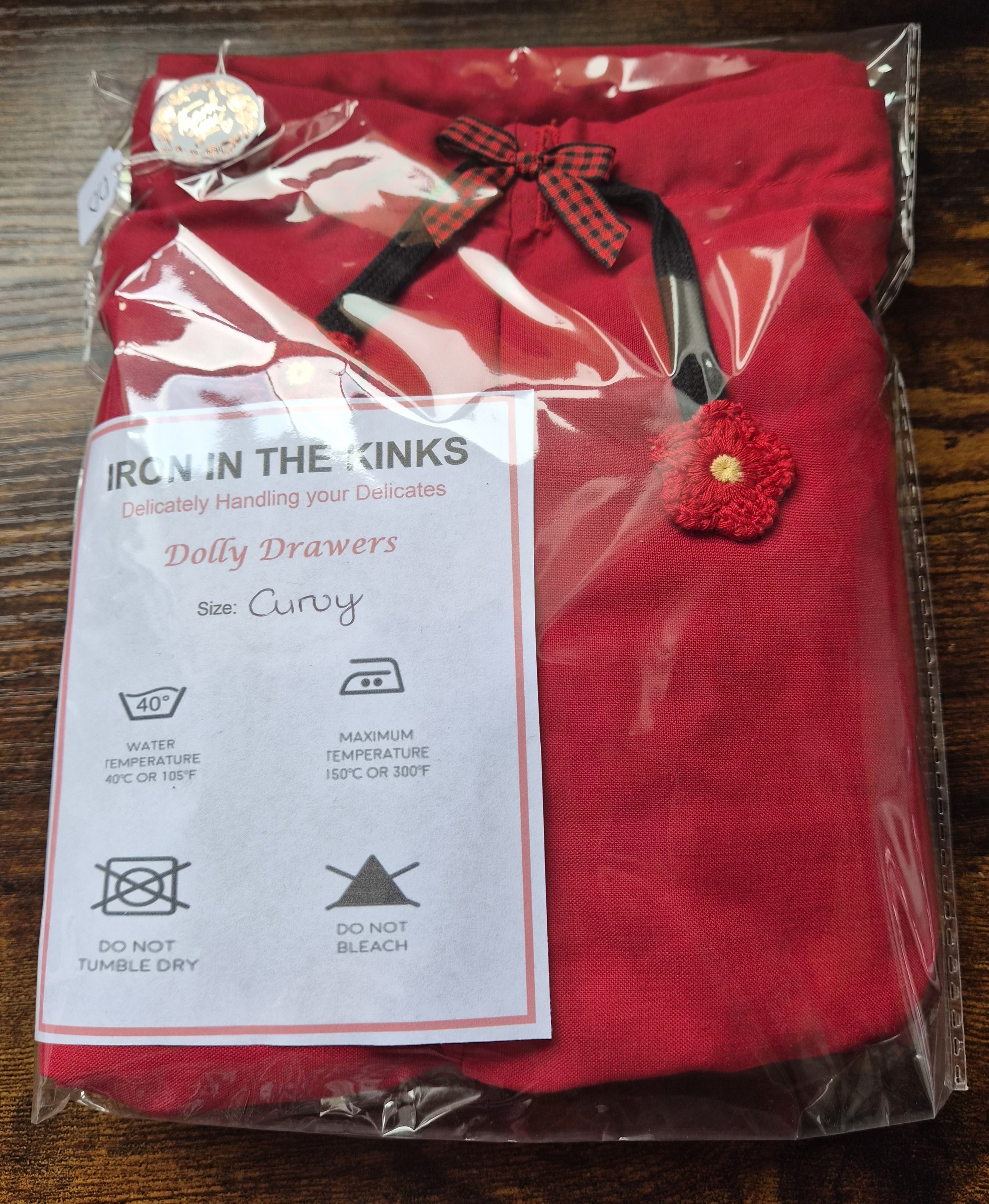 A folded red short in clear plastic packaging, featuring a red garmentwith a black and red gingham bow, a red flower applique with a gold center, and a white care instruction card. sissy knickers, adult baby nappy cover, victorian style drawers, unisex