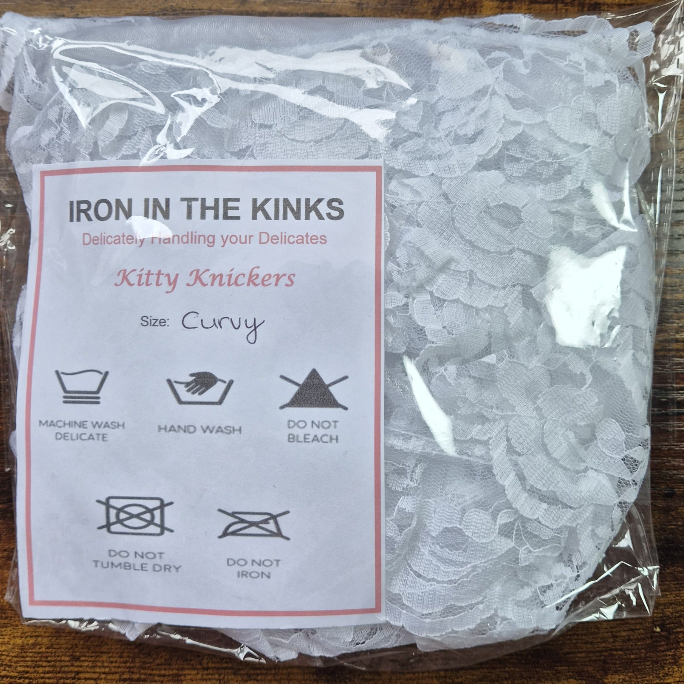 Iron in the Kinks Kitty Knickers in white lace in clear packaging with care label, size Curvy — machine wash delicate, hand wash, do not bleach, tumble dry, or iron