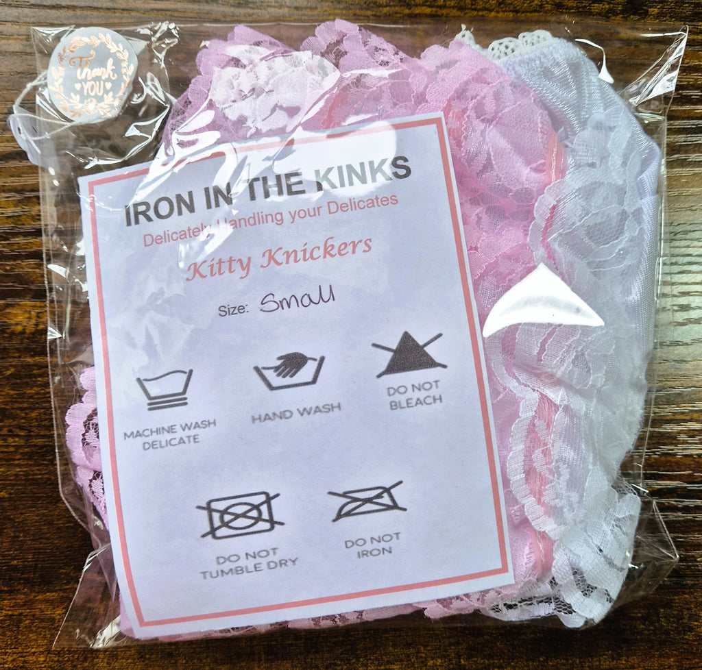 Pink and white lace 'Kitty Knickers' in a clear plastic bag with a white care label that reads 'IRON IN THE KINKS' and shows washing and ironing instructions, placed on a wooden surface.