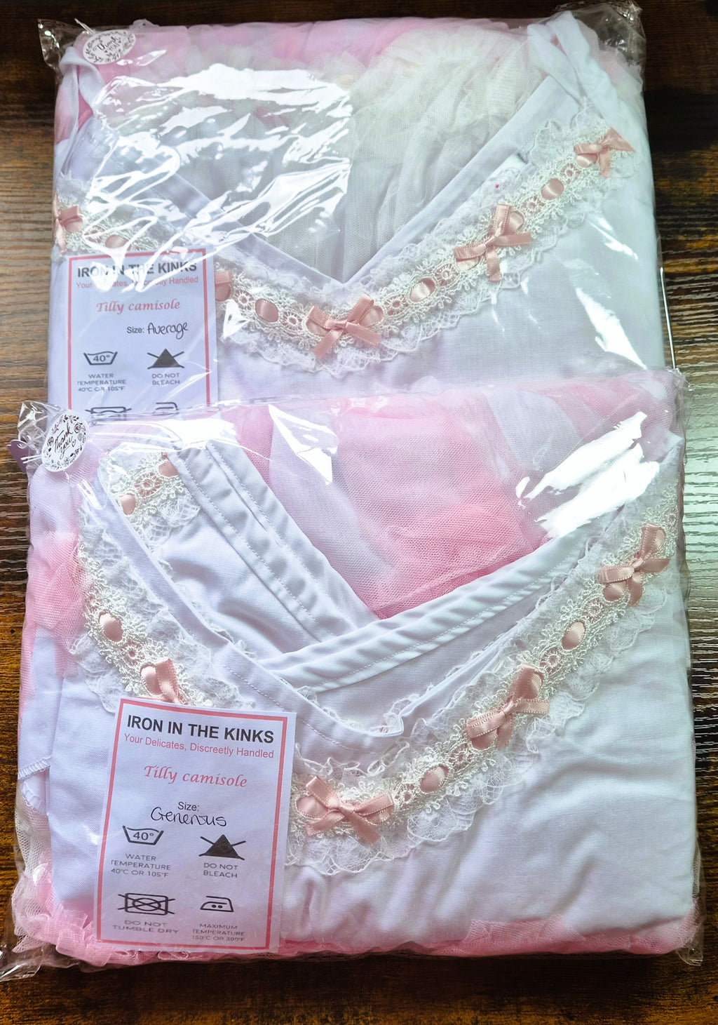 Two Iron in the Kinks Tilly Camisoles in clear packaging — Average and Generous sizes — showing white fabric with broderie anglaise V-neckline and pink ribbon bows