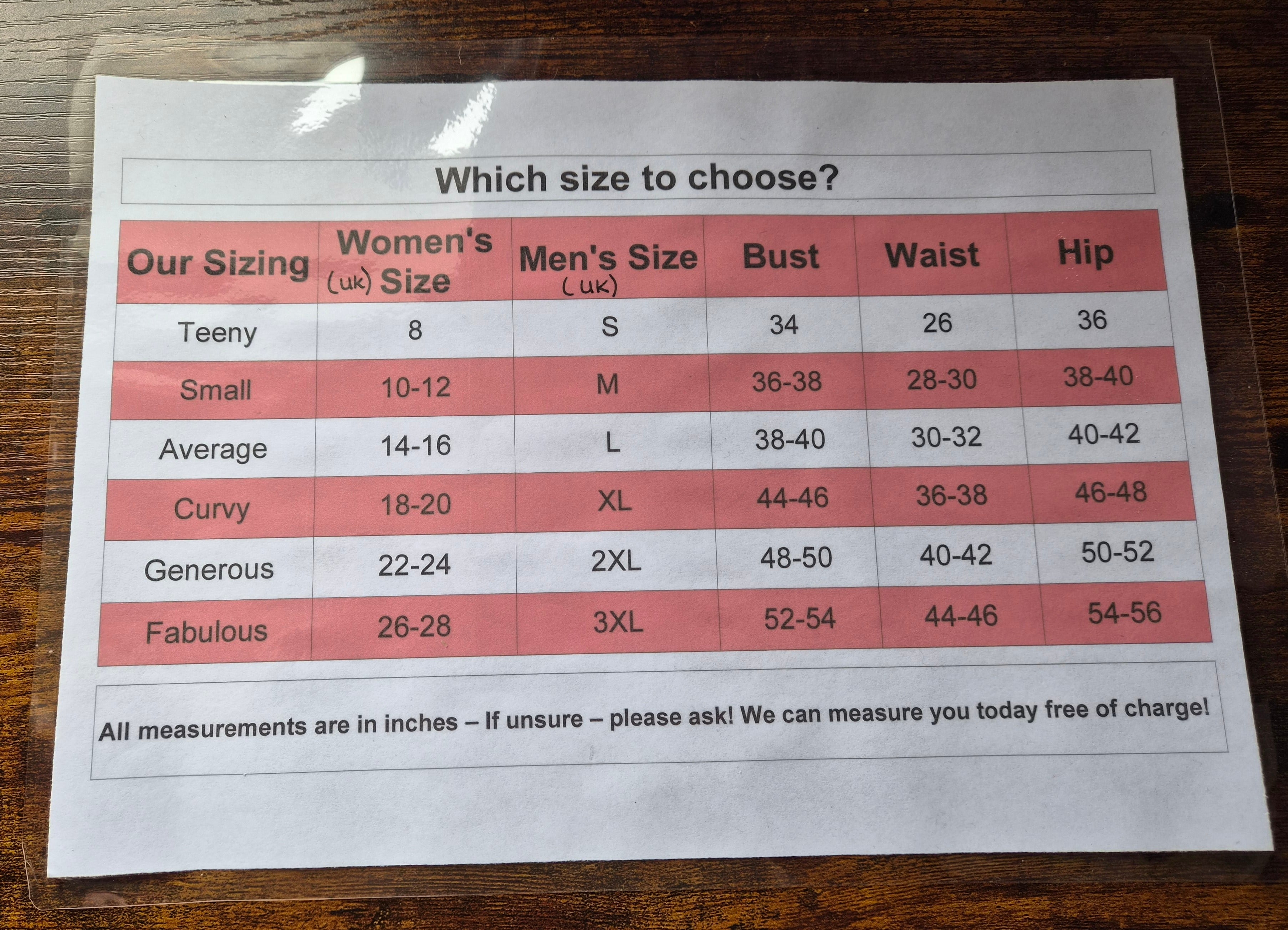 A printed sizing chart titled 'Which size to choose?' showing a size guide table with columns for Our Sizing, Women's (UK) Size, Men's Size (UK), Bust, Waist, and Hip, placed on a dark wooden surface.