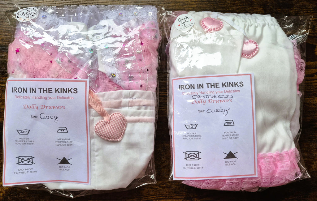 Two packaged sets of drawers by Iron in the Kinks, one set in pink and white and the other in white with pink accents, displayed side-by-side on a dark wooden surface. sissy knickers, adult baby nappy cover, victorian style drawers, unisex