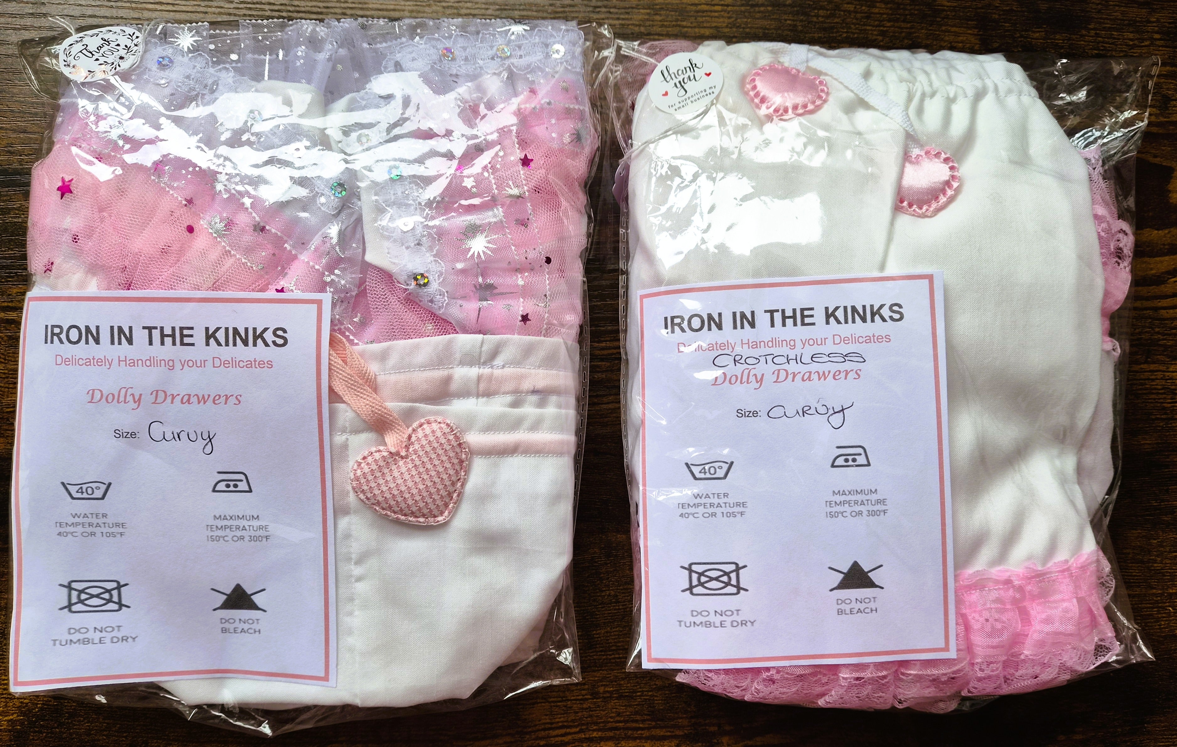 Two packaged sets of drawers by Iron in the Kinks, one set in pink and white and the other in white with pink accents, displayed side-by-side on a dark wooden surface. sissy knickers, adult baby nappy cover, victorian style drawers, unisex