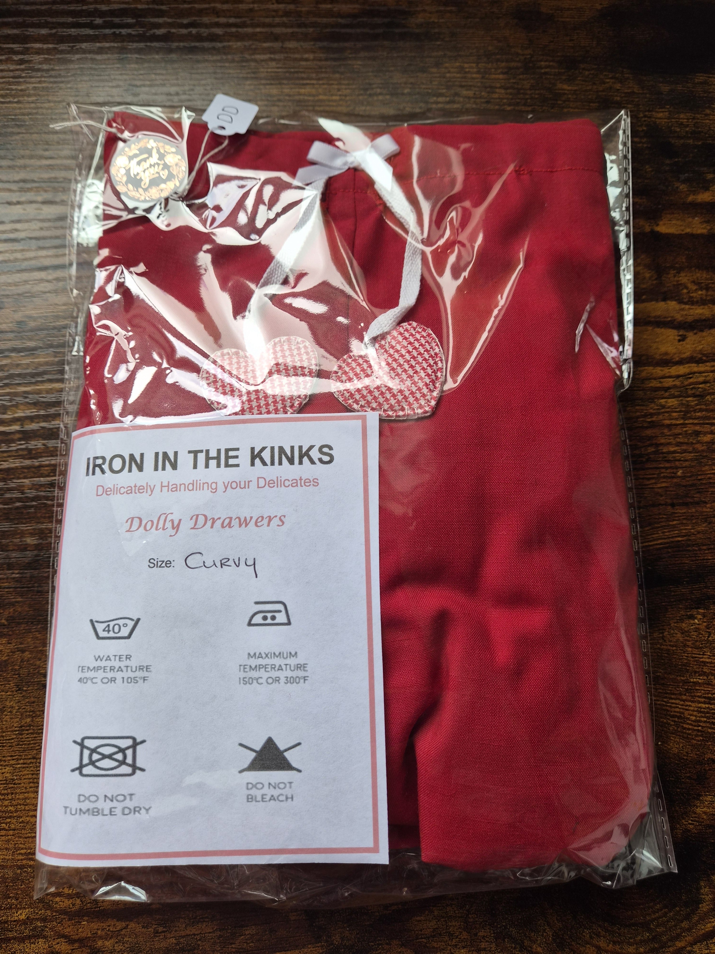 A folded red garment package sealed in clear plastic, with a white care label reading 'IRON IN THE KINKS' and 'Dolly Drawers' on a dark wooden surface. sissy knickers, adult baby nappy cover, victorian style drawers, unisex