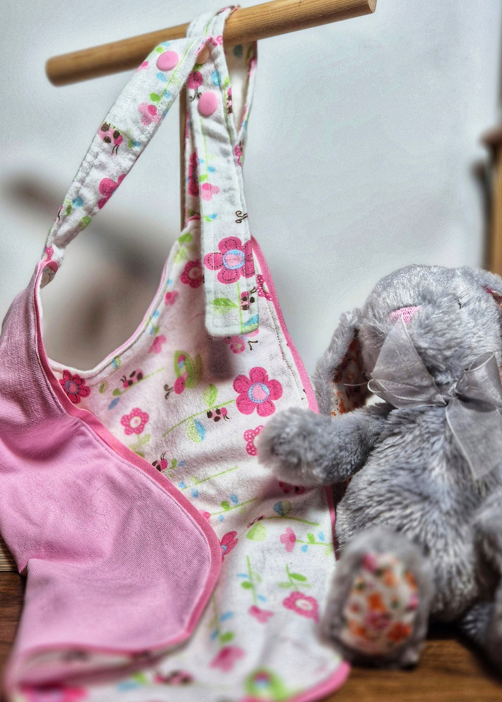 Floral-patterned adult baby bib with pink accents on a wooden hanger next to a gray stuffed rabbit toy.