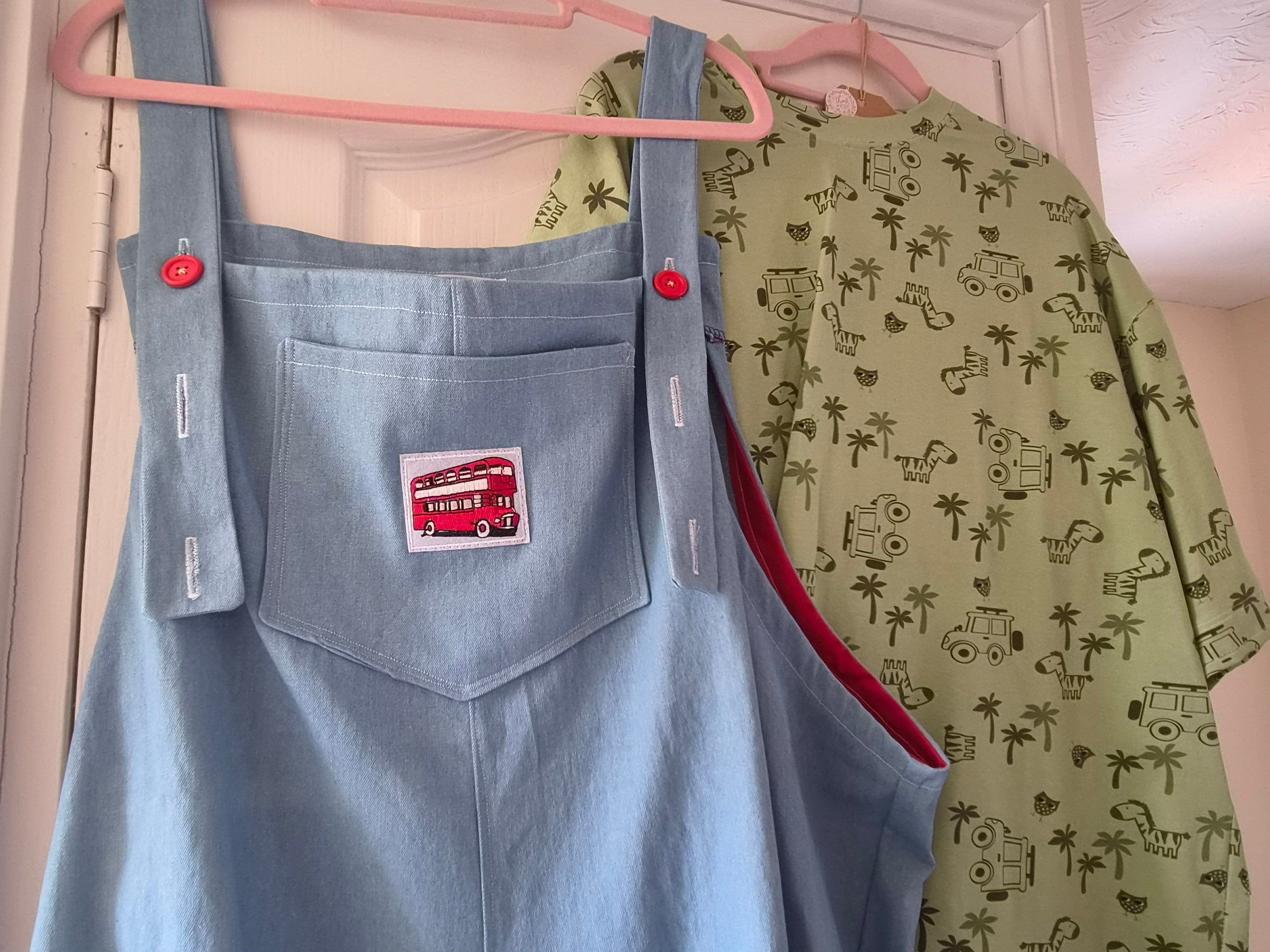 A light blue denim overall-style bib with a front pocket and a red double-decker bus patch, hanging on a pink hanger next to a green short-sleeve shirt with a palm tree and vehicle print.