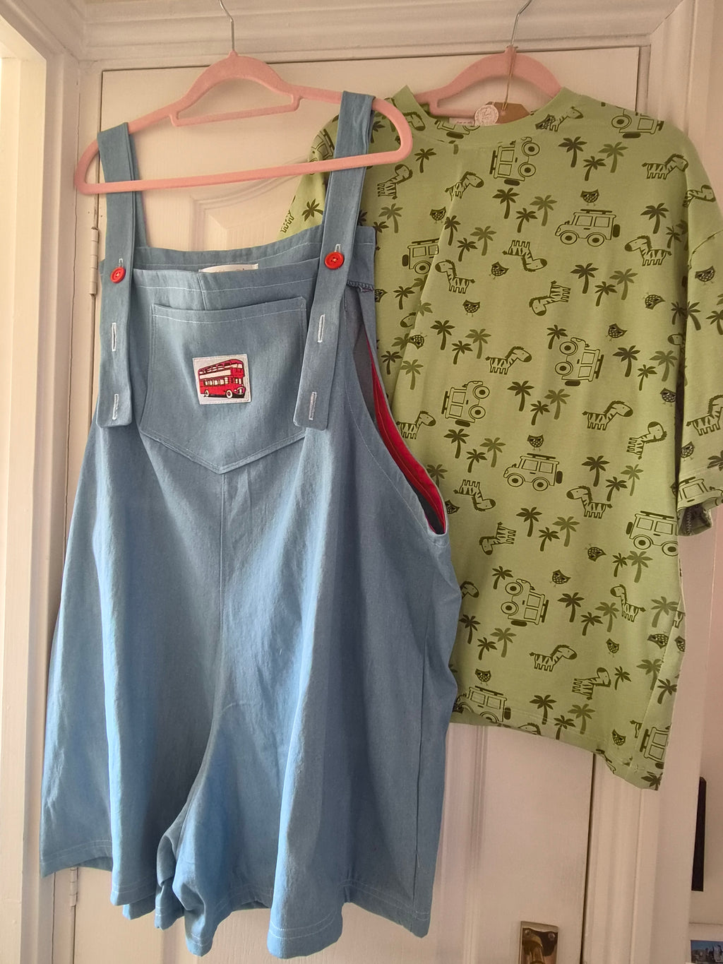 A light blue denim overall shortee laid flat on a pink hanger, with a green patterned shirt partially visible behind it on a white door.