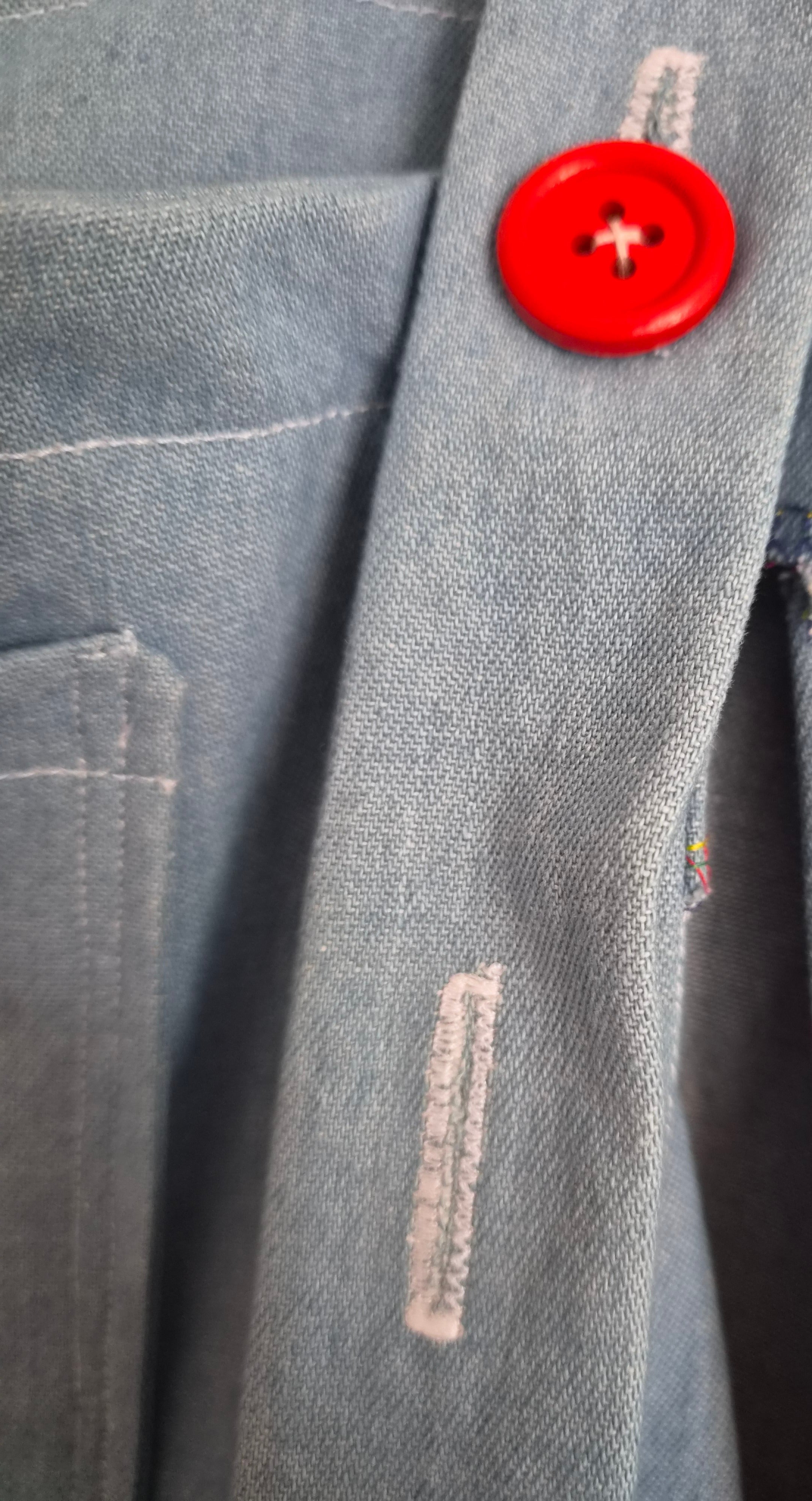 Close-up of a light blue denim garment featuring a prominent bright red button and white contrast stitching.