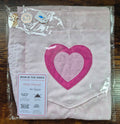 A folded cotton playsuit in light pink fabric with a large pink heart applique, sealed in clear plastic packaging with a product tag and care label visible.