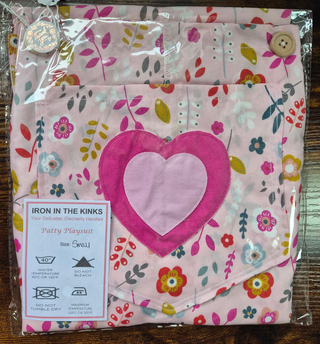 Iron in the Kinks Patty Playsuit in white with pink appliqué double heart motif in clear packaging with care label, size Small — wash at 40°C, do not bleach, do not tumble dry, iron max 150°C