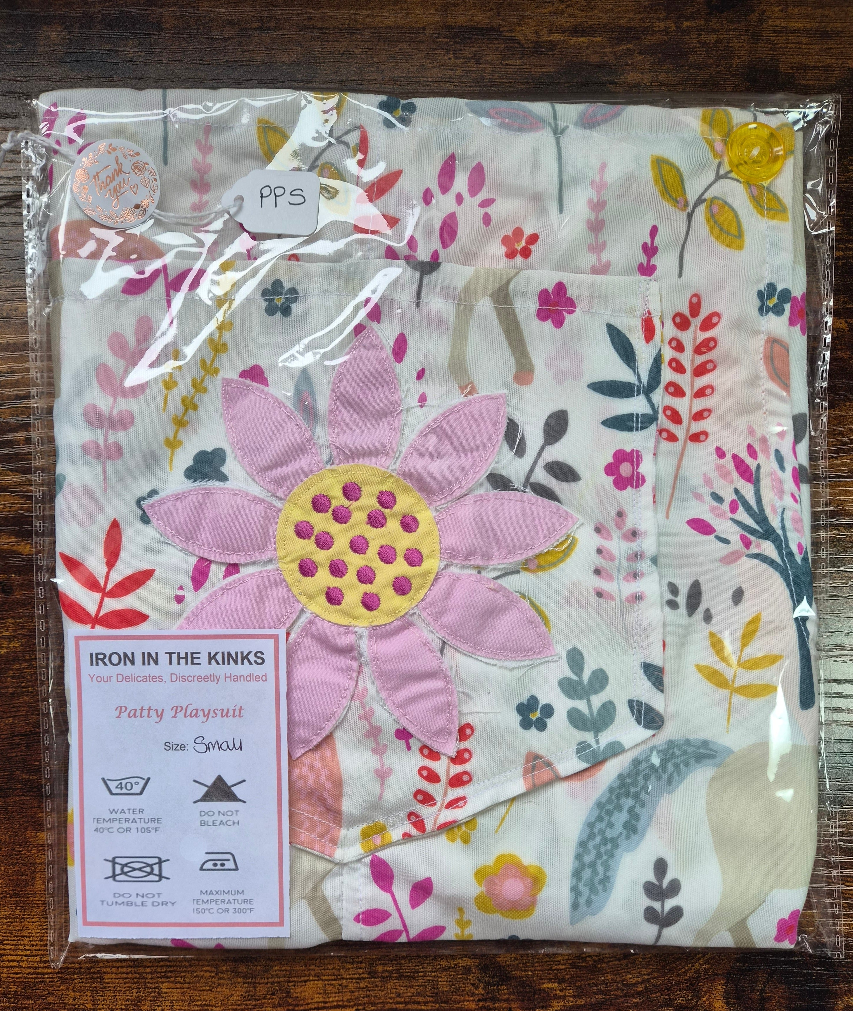 Iron in the Kinks Patty Playsuit in white  unicorn floral print with pink appliqué double daisy  motif in clear packaging with care label, size Small — wash at 40°C, do not bleach, do not tumble dry, iron max 150°C
