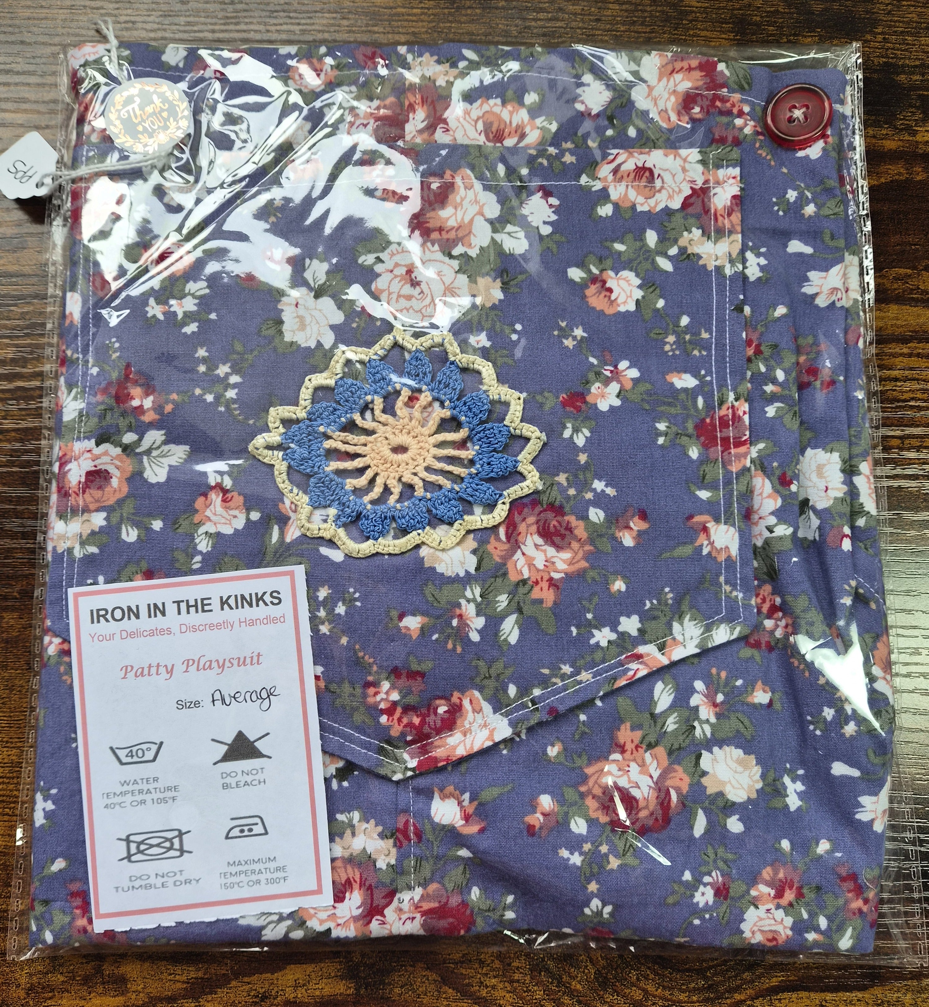 Iron in the Kinks Patty Playsuit in blue vintage floral print in clear packaging with crochet mandala embellishment and care label, size Average — wash at 40°C, do not bleach, do not tumble dry, iron max 150°C