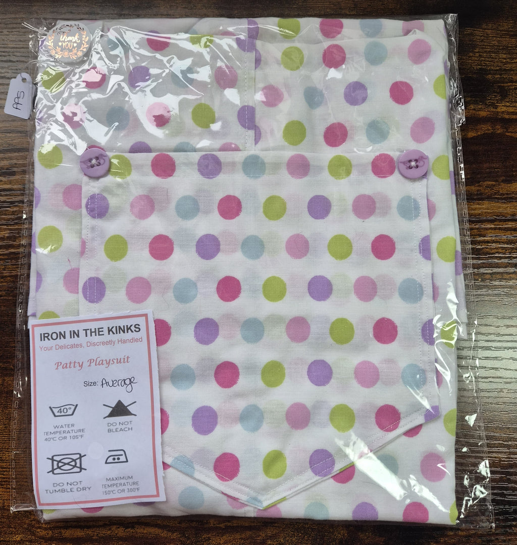 A packaged cotton  play suit with a colorful polka dot pattern, featuring a buttoned bib and an attached care label that reads 'Iron in the Kinks'.