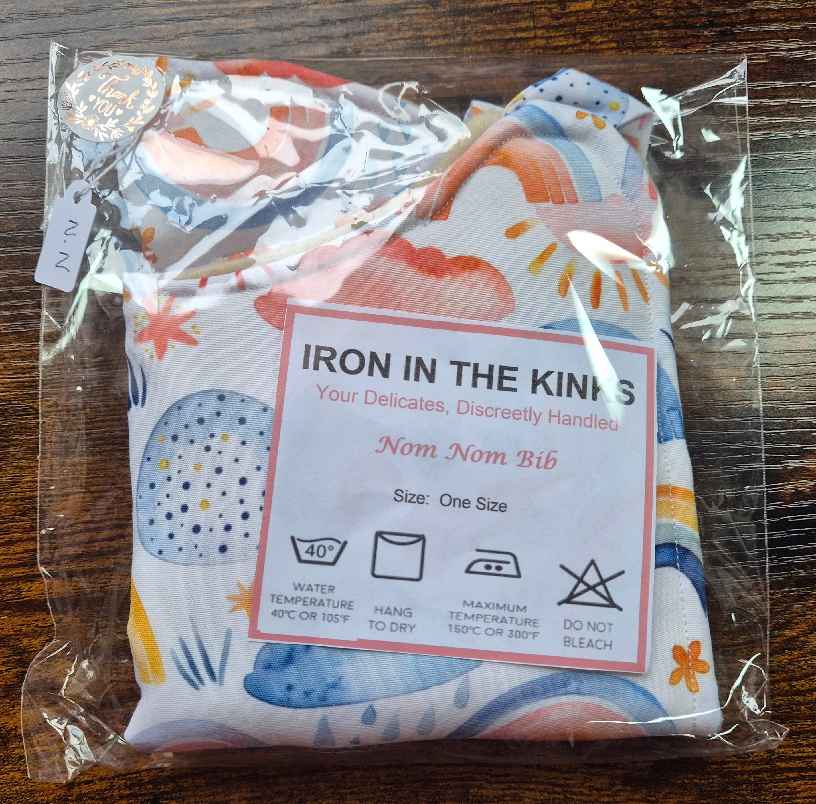 Iron in the Kinks Nom Nom Bib in rainbow and cloud print in clear packaging with care label — wash at 40°C, hang to dry, iron max 150°C, do not bleach, one size