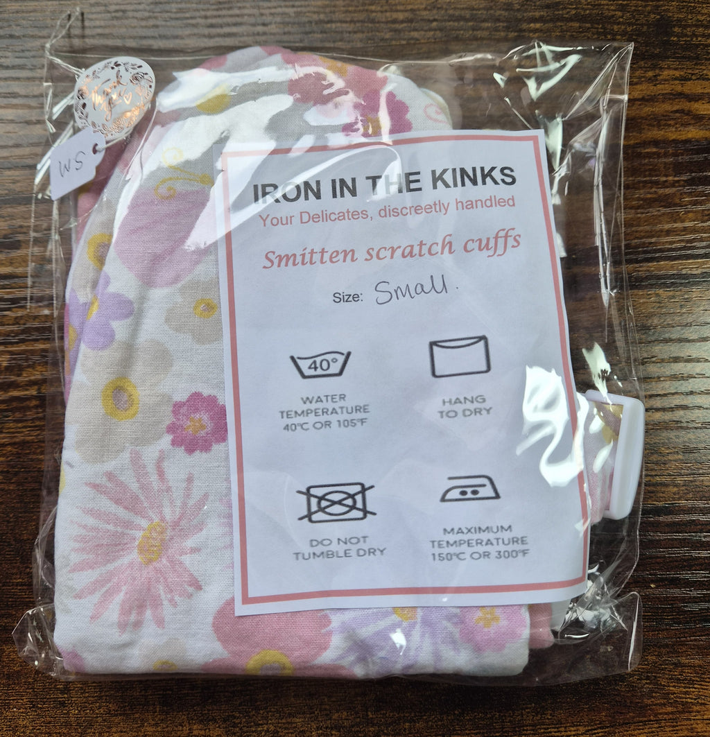 A packaged set of small smitten scratch cuffs with a white instruction card reading 'IRON IN THE KINKS' and 'Smitten scratch cuffs Size Small,' photographed on a dark wooden surface.