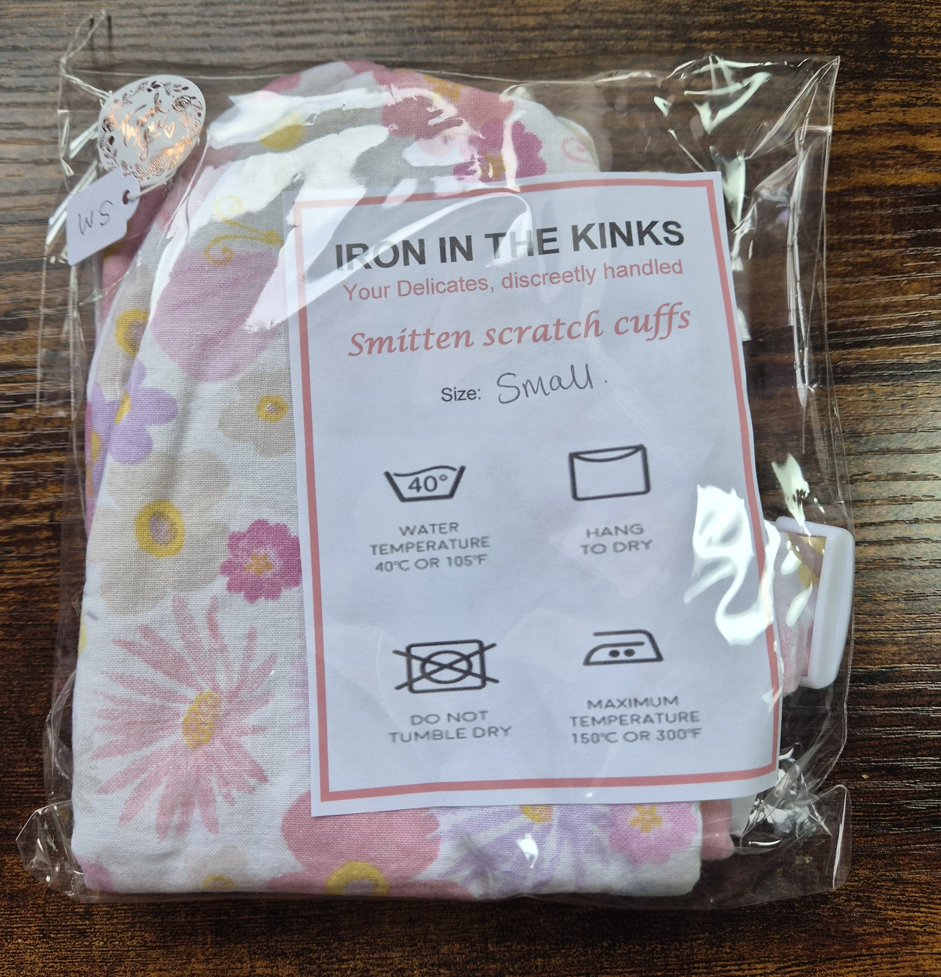 A packaged set of small smitten scratch cuffs with a white instruction card reading 'IRON IN THE KINKS' and 'Smitten scratch cuffs Size Small,' photographed on a dark wooden surface.