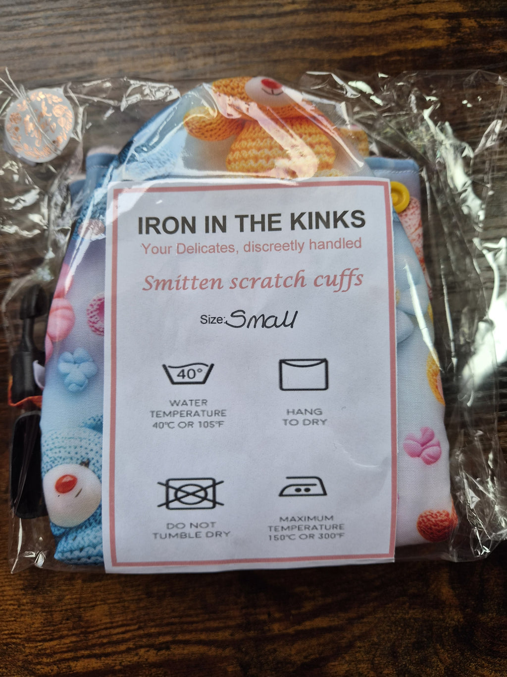 A packaged set of small smitten scratch cuffs with a white instruction card reading 'IRON IN THE KINKS' and 'Smitten scratch cuffs Size Small,' photographed on a dark wooden surface.