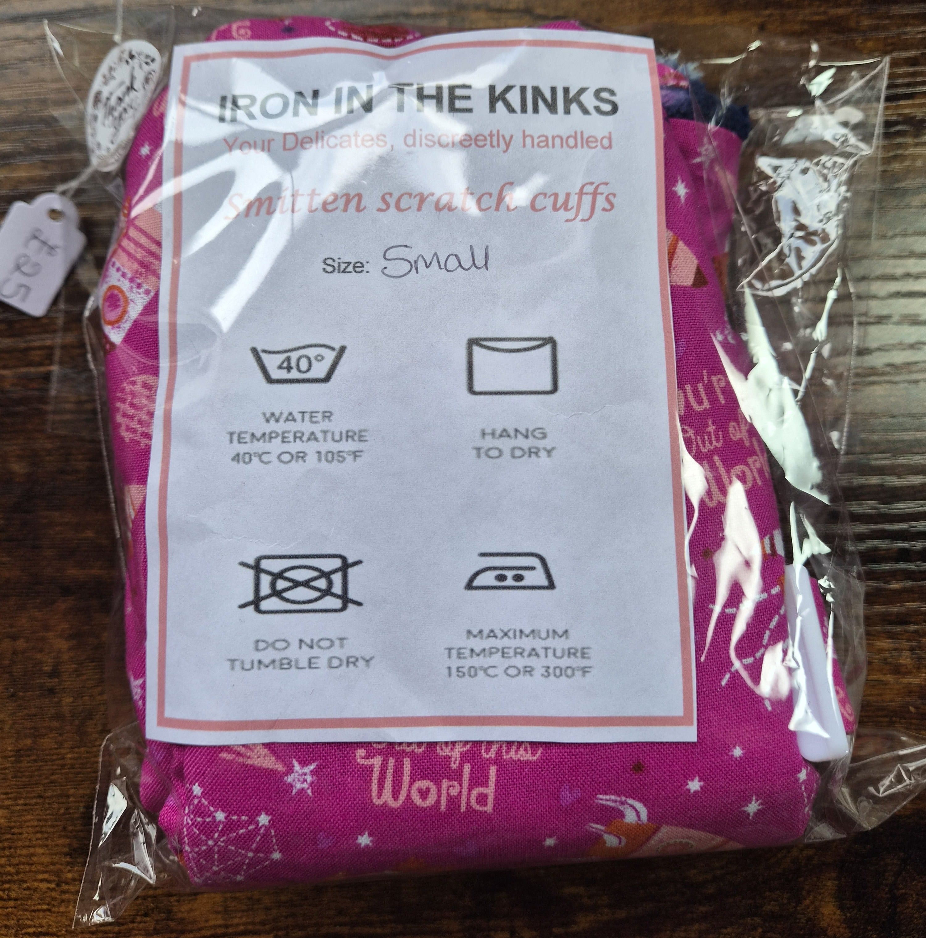 A packaged set of small smitten scratch cuffs with a white instruction card reading 'IRON IN THE KINKS' and 'Smitten scratch cuffs Size Small,' photographed on a dark wooden surface.