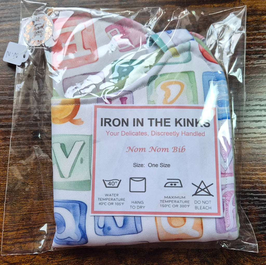 Iron in the Kinks Nom Nom Bib in alphabet blocks print in clear packaging with care label — wash at 40°C, hang to dry, iron max 150°C, do not bleach, one size