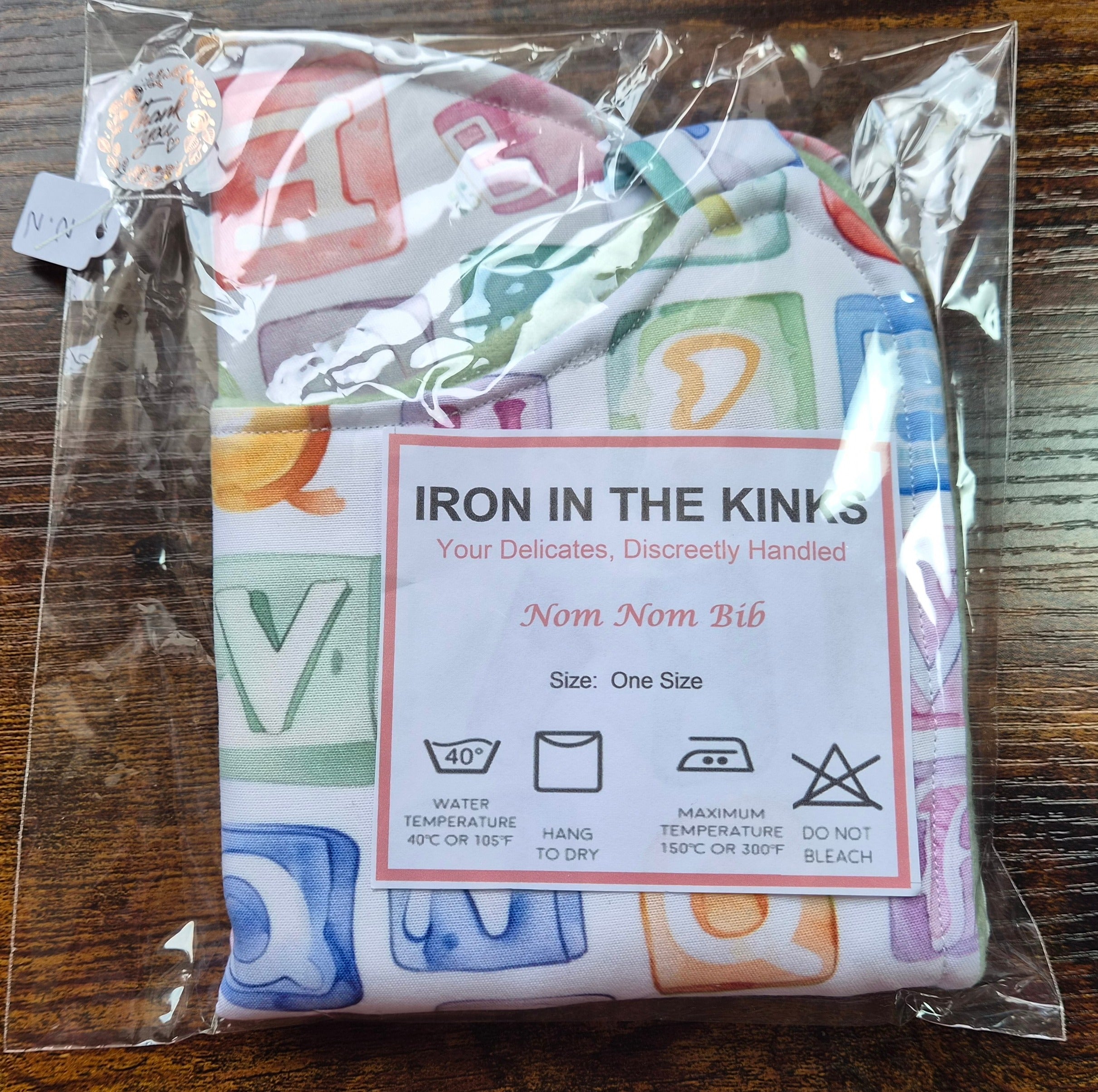 Iron in the Kinks Nom Nom Bib in alphabet blocks print in clear packaging with care label — wash at 40°C, hang to dry, iron max 150°C, do not bleach, one size