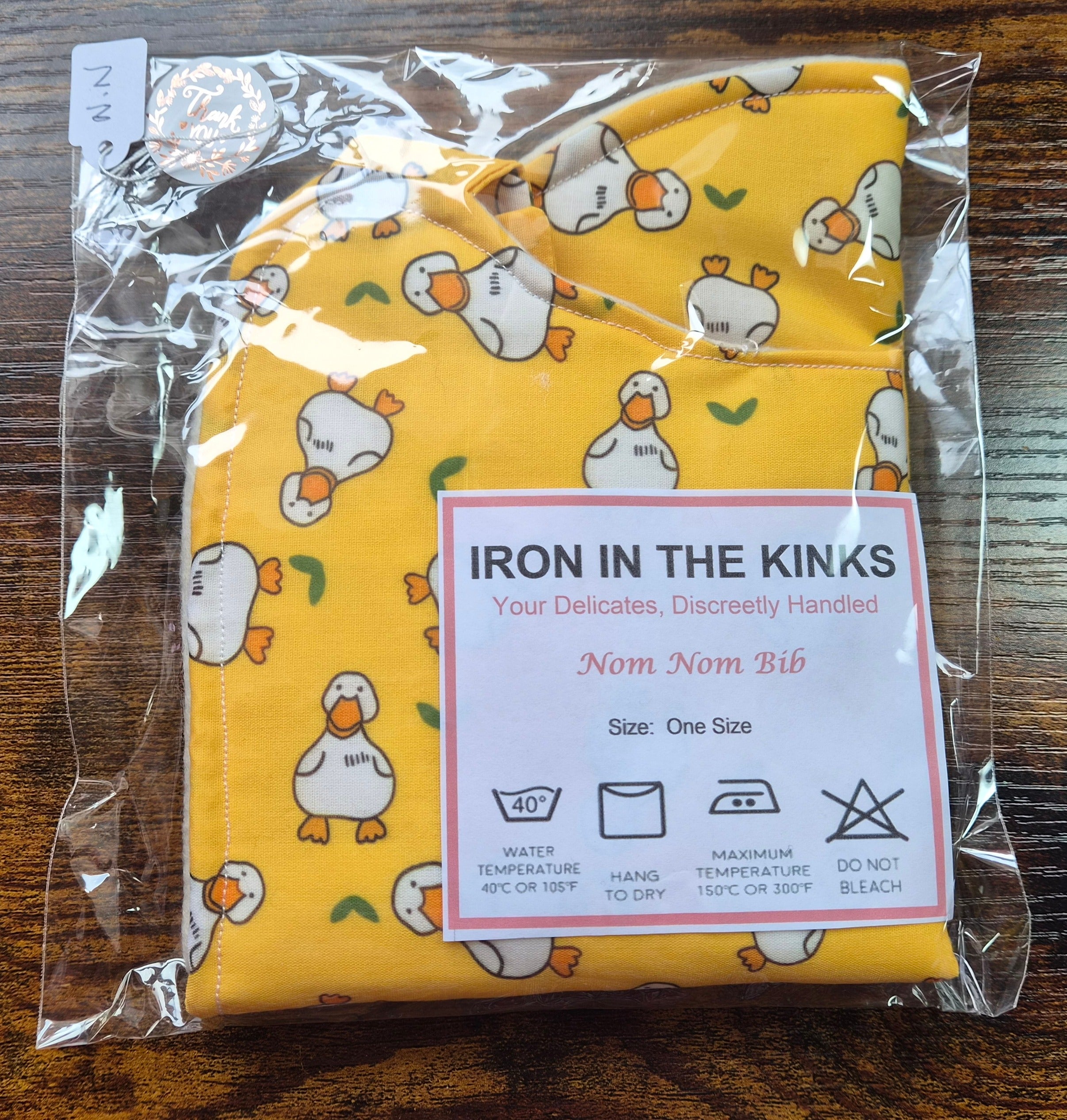 Iron in the Kinks Nom Nom Adult Bib in yellow chunky duck in clear packaging with care label — wash at 40°C, hang to dry, do not bleach, one size