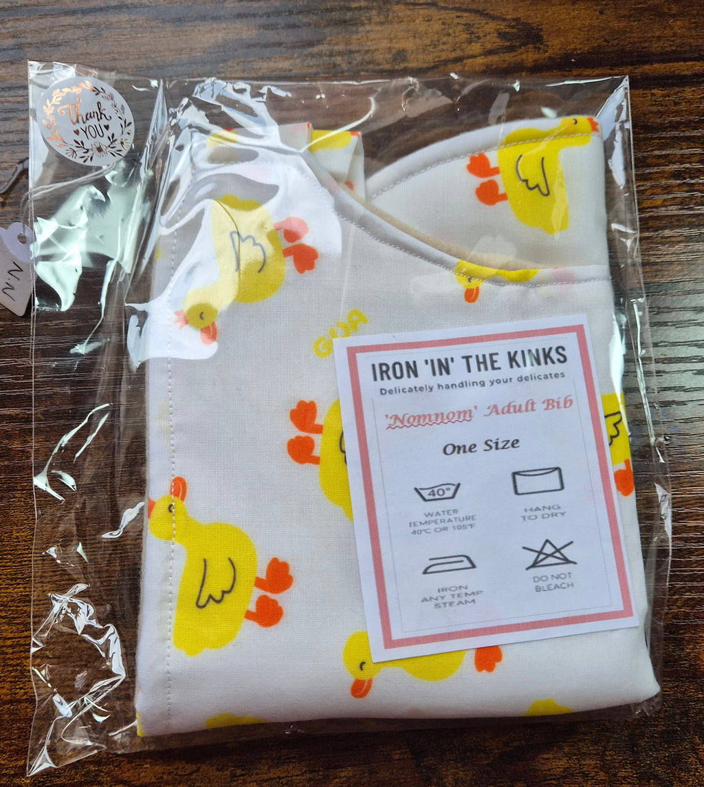 Iron in the Kinks Nom Nom Bib in yellow duck print in clear packaging with care label — wash at 40°C, hang to dry, iron max 150°C, do not bleach, one size