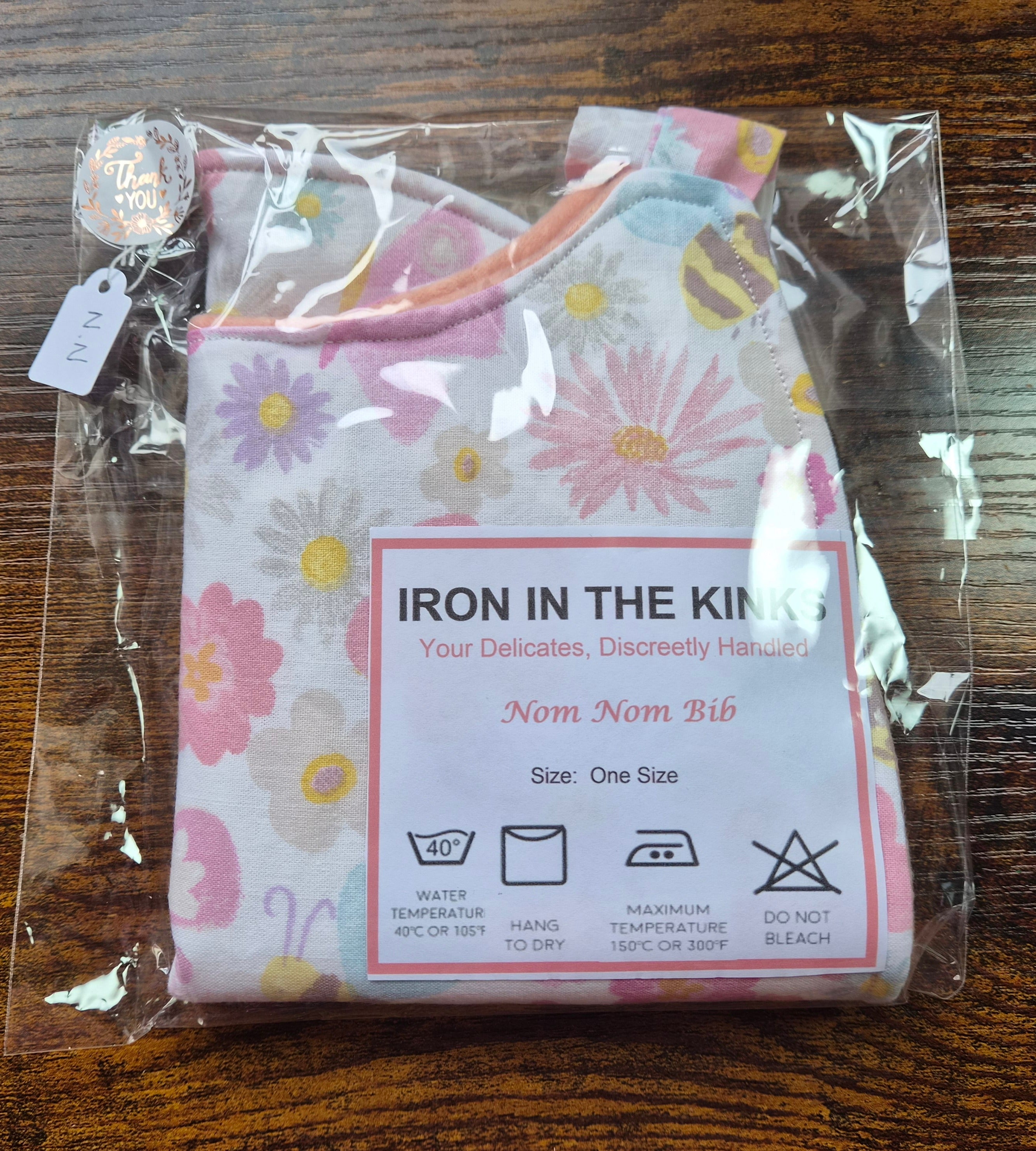 Iron in the Kinks Nom Nom Bib in pink daisy and bee print in clear packaging with care label — wash at 40°C, hang to dry, iron max 150°C, do not bleach, one size