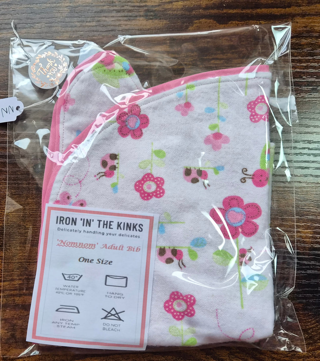Iron in the Kinks Nom Nom Adult Bib in pink floral and ladybird print with pink binding in clear packaging with care label — wash at 40°C, hang to dry, do not bleach, one size