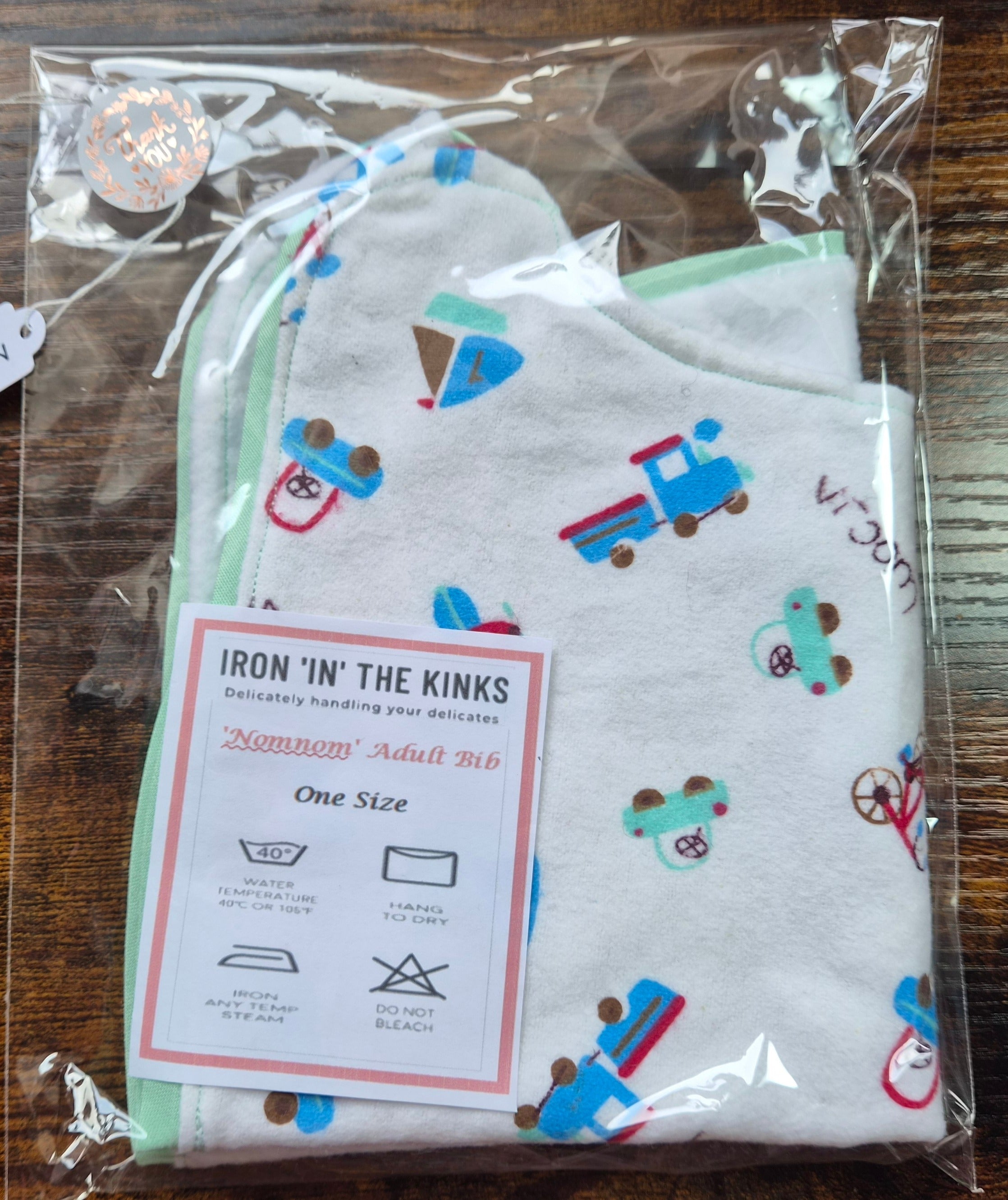 Iron in the Kinks Nom Nom Adult Bib in white toy trains and vehicles print with mint green binding in clear packaging with care label — wash at 40°C, hang to dry, do not bleach, one size