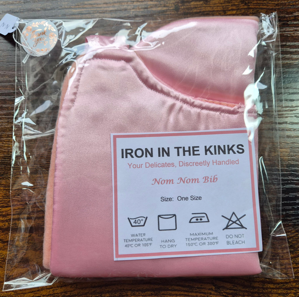 Iron in the Kinks Nom Nom Bib in plain pink satin in clear packaging with care label — wash at 40°C, hang to dry, iron max 150°C, do not bleach, one size