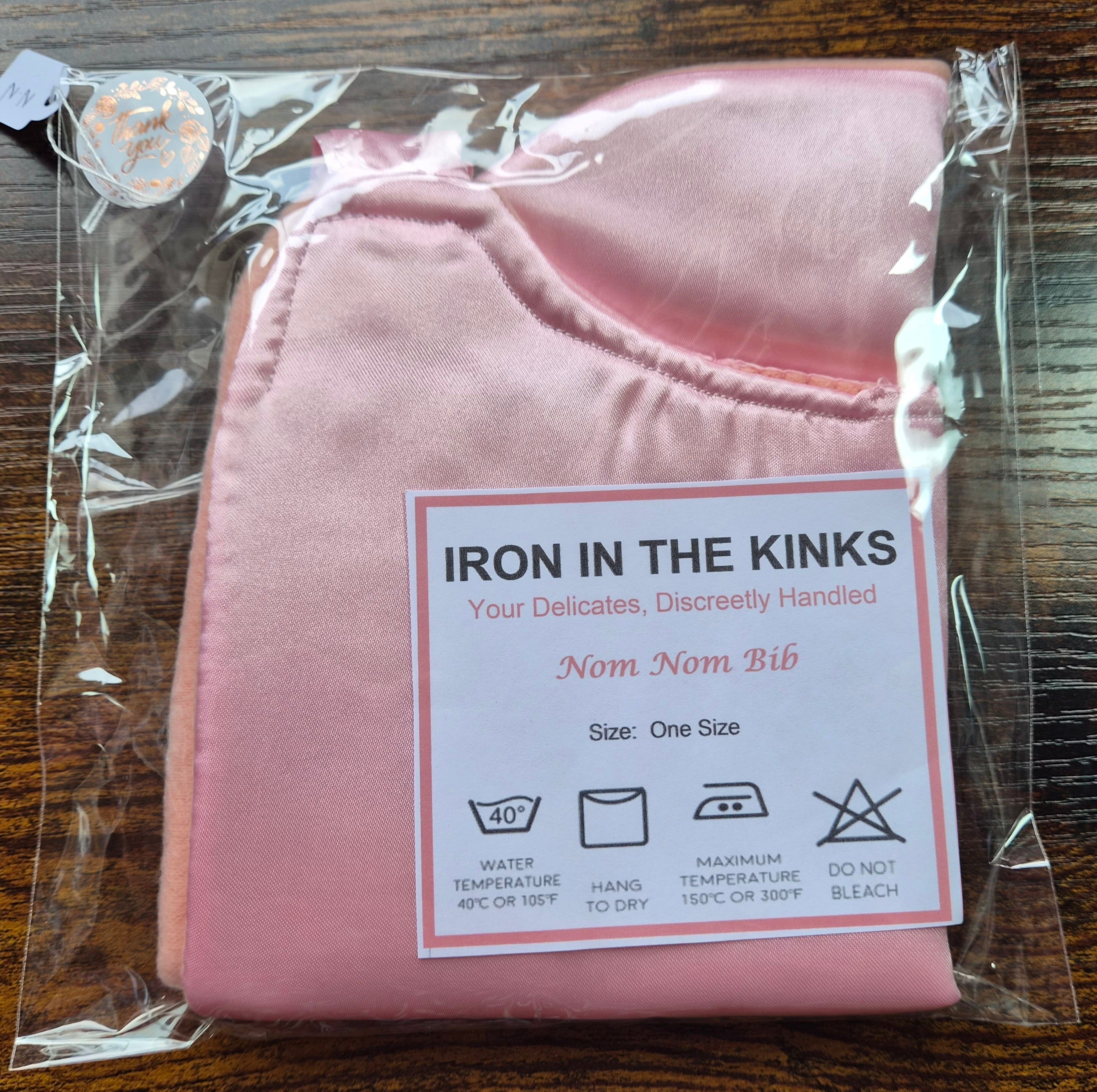 Iron in the Kinks Nom Nom Bib in plain pink satin in clear packaging with care label — wash at 40°C, hang to dry, iron max 150°C, do not bleach, one size