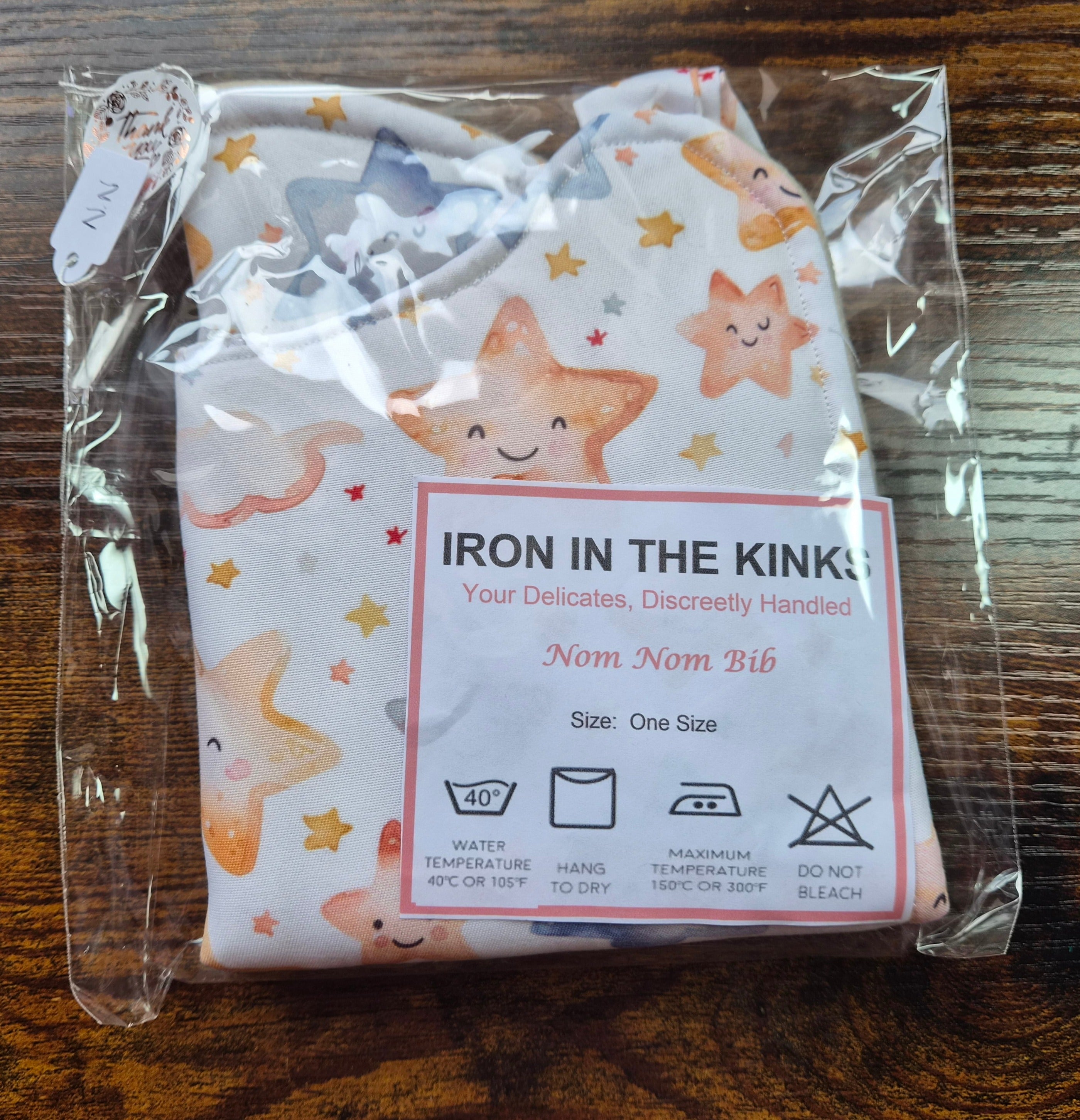 Iron in the Kinks Nom Nom Bib in smiling stars print in clear packaging with care label — wash at 40°C, hang to dry, iron max 150°C, do not bleach, one size